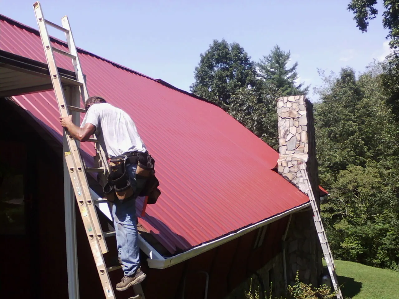Roofer on a ladder beside a red metal roof and stone chimney — Soffit & Fascia Repair by Roofing Brothers in Monongahela